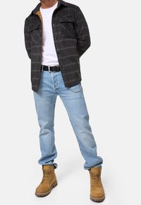Sort ternet flannel skjorte med to brystlommer over en hvid t-shirt, parret med lyseblå jeans og brune arbejdssko. Har sølv kædedetalje.