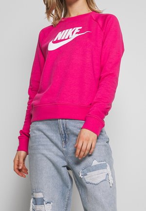 Personne portant un sweat-shirt Nike rose vif avec un logo blanc et un jean bleu clair déchiré, debout devant un arrière-plan gris uni.