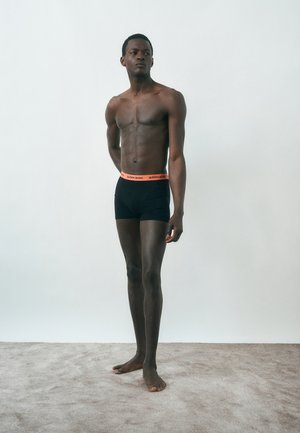 Ψηλός άνδρας, όρθιος ξυπόλητος σε χαλί, φορούμενος μαύρα μπόξερ με πορτοκαλί μέση με την επιγραφή «BJÖRN BORG», κοιτάζει στο πλάι σε απλό φόντο.