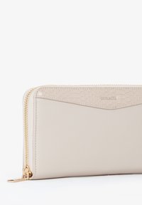 Portafoglio beige in pelle traforata con finitura liscia. Presenta una cerniera dorata e un logo in rilievo. Design geometrico lungo la parte superiore.