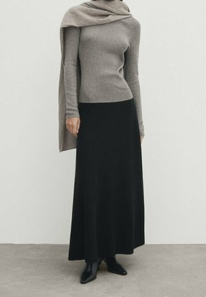Personne portant un haut à manches longues taupe avec un châle assorti, une longue jupe noire et des bottines noires, debout devant un fond uni.