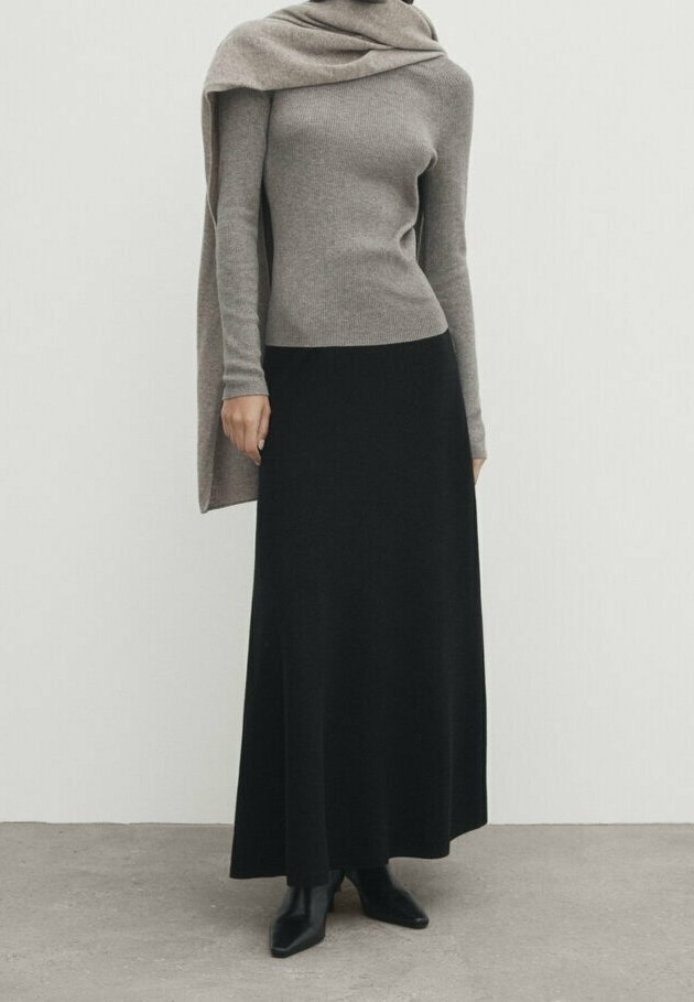 Personne portant un haut à manches longues taupe avec un châle assorti, une longue jupe noire et des bottines noires, debout devant un fond uni.
