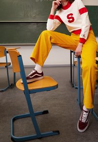 Bílá mikina s červenými akcenty a logem „S“, spárovaná se žlutými kordovým širokými kalhotami. Burgundské tenisky s bílými tkaničkami.