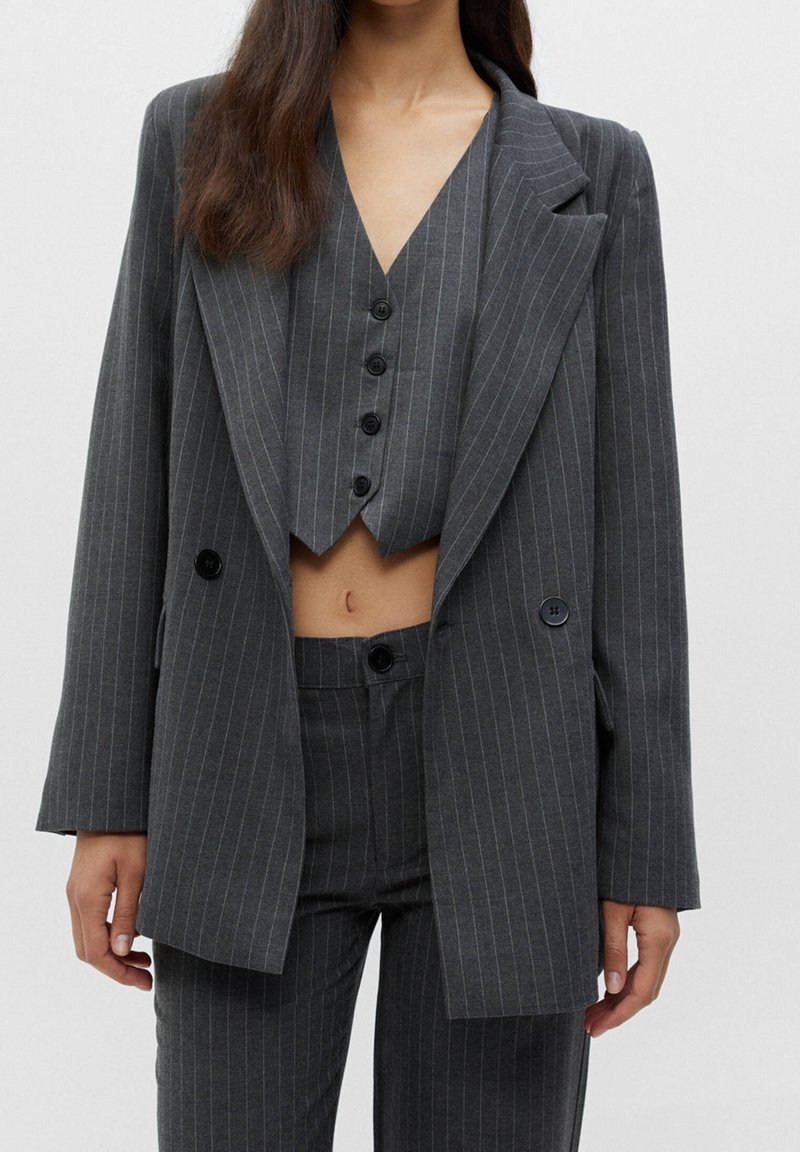 Femme portant un costume gris foncé à fines rayures avec un blazer croisé, un gilet court assorti et un pantalon taille haute.