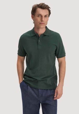 Junger Mann trägt ein dunkelgrünes kurzärmeliges Poloshirt und blaue Hose, steht mit einer Hand in der Tasche vor einfachem Hintergrund.