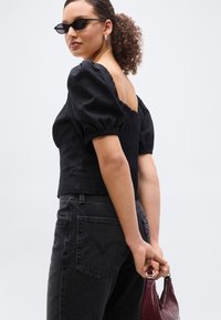 Femme aux cheveux bouclés et lunettes de soleil, portant un haut noir à manches bouffantes et un jean foncé, tenant un sac à main bordeaux derrière son dos.