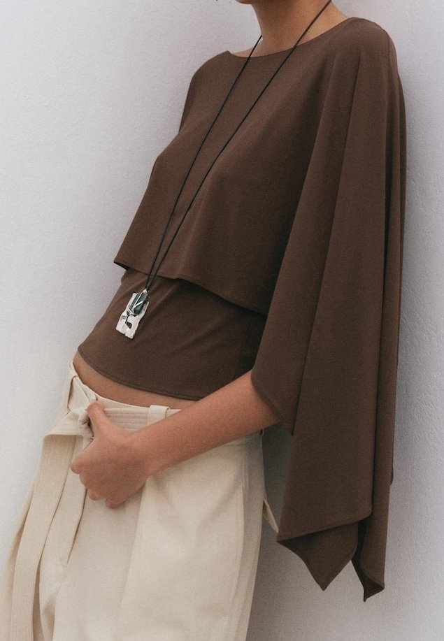 Femme portant un haut marron ample à manches larges et un long collier pendentif, ajustant un pantalon crème taille haute avec une ceinture nouée.