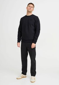 Zwarte katoenen sweatshirt met ronde hals en getextureerd logo, gecombineerd met zwarte broeken en witte sneakers met bruine accenten.
