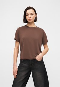 Νεαρό άτομο με κοντά σκούρα μαλλιά, φορώντας καφέ κοντομάνικο πουκάμισο και μαύρο τζιν με φαρδιά πόδια, στέκεται με το ένα χέρι στην τσέπη.