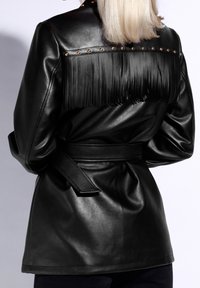 Chaqueta de cuero negro con detalle de flecos en la parte posterior, que presenta acentos dorados en los hombros y un cinturón anudado en la cintura. Textura suave.