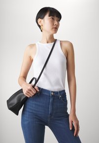 Débardeur blanc à côtes assorti avec un jean taille haute bleu foncé. Le modèle tient un sac à main noir avec une bandoulière courte. Design simple, sans motifs.