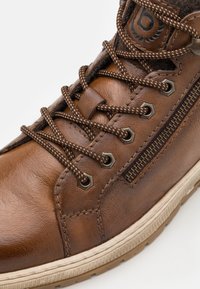 Brun läder högskosko med texturerad yta, kontrasterande bruna snörningar, metallöglor, sidodragkedja och gummisula med beige accent.