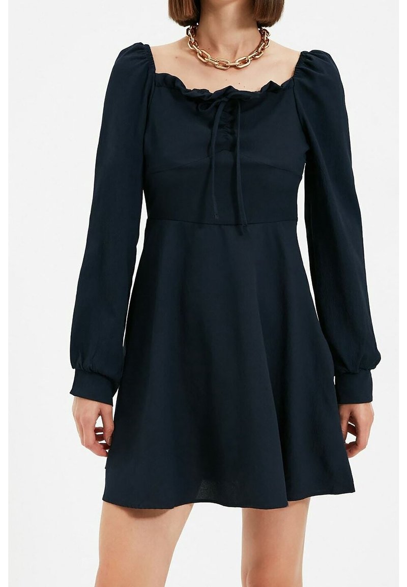 Femme portant une robe noire à manches longues avec un décolleté carré et un détail volanté, associée à un collier chaîne doré épais.