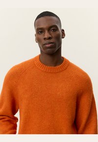 Giovane uomo con pelle scura che indossa un maglione arancione lavorato a maglia, rivolto in avanti con un'espressione neutra su uno sfondo semplice.