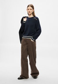 Πλεκτό μπλε ναυτικό πουλόβερ με μανσέτες και τελείωμα με λευκές ρίγες, τρία χρυσά κουμπιά στον ώμο, συνδυασμένο με άνετο καφέ παντελόνι κορντόι και μαύρα παπούτσια.