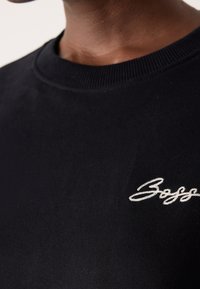 Gros plan d'un t-shirt noir col rond avec le mot blanc brodé « Boss » sur la poitrine, porté par une personne à la peau foncée.