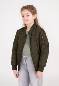 Olivengroene bomberjack met een ribgebreide kraag, een rits aan de voorkant en een zak op de mouw; gecombineerd met een ribgebreid topje en lichtgrijze jeans.