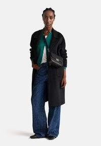 Cappotto di lana nera su un cardigan verde, camicia bianca, jeans a gamba larga e scarpe nere; tiene in mano una piccola borsa a tracolla a righe.