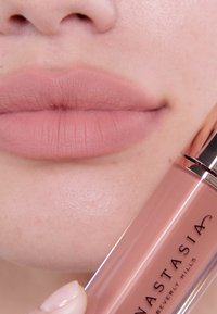 Matowy, nagi błyszczyk trzymany obok gładkich ust. Błyszczyk w przezroczystej tubie z metalicznymi różowo-złotymi akcentami i logo marki. Usta pokazują naturalną teksturę.