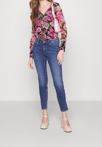 Top floreale a maniche lunghe in nero, rosa e verde; abbinato a jeans aderenti blu e tacchi a pianta beige con cinturini alla caviglia.
