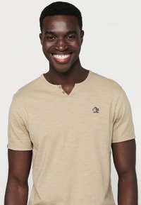 Beige, rövid ujjú póló henley nyakkal, két gombbal és egy kicsi fekete logóval a mellkasán. Sima textúra.