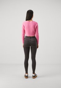 Ženska stoji obrnjena stran, oblečena v roza kratko pulover, temno sivo oprijete kavbojke in črno-bel športni copate z gumijastimi podplati.