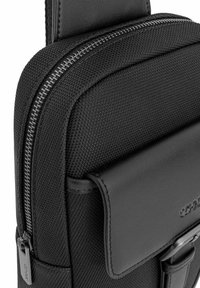 Czarna torba nylonowa z teksturowanym wykończeniem, kontrastującymi skórzanymi akcentami, zapięciem na zamek oraz regulowanym paskiem do łatwego noszenia.