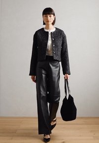 Veste en tweed noir avec des boutons argentés, blouse blanche et pantalons larges en cuir noir. Porte un sac fourre-tout noir et des ballerines noires à bout pointu.