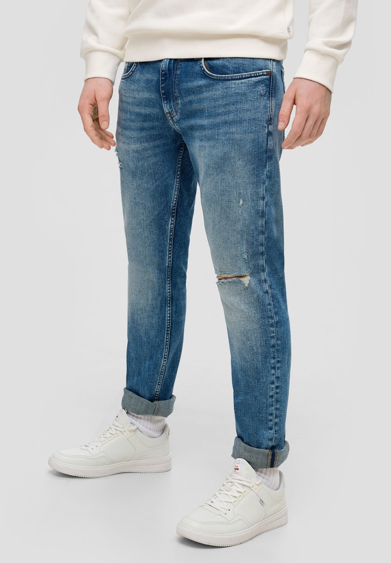 Blauwe denim jeans met een slim fit, versleten details en omgeslagen broekspijpen, gecombineerd met witte sneakers en witte crew-sokken.
