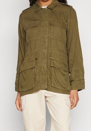 Kvinde iført olivengrøn utility-jakke med fire frontlommer med klap og cremefarvede cargobukser, stående mod en ensfarvet baggrund.