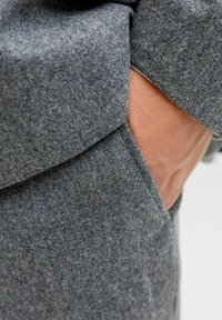 Szary filcowy materiał o gładkiej fakturze, z boczną kieszenią i subtelnym detalem szwu. Zbliżenie na materiał i ułożenie dłoni.