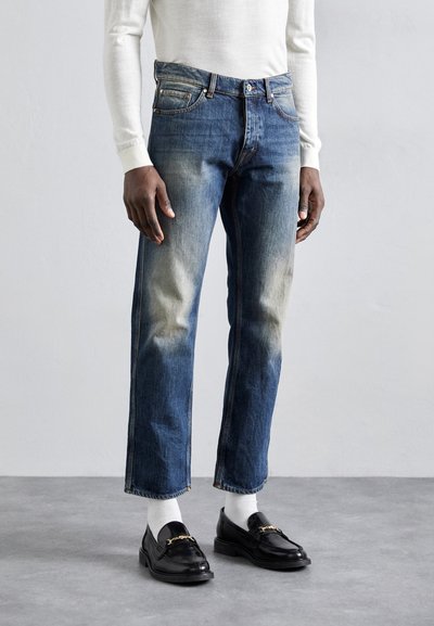 Blaue Jeans aus Denim mit einem leicht verblassten Waschung und geradem Schnitt, kombiniert mit schwarzen Slippern und weißen Socken. Einfaches Design mit minimalem Zubehör.