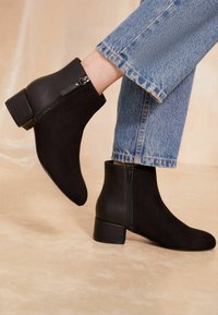 Persona que lleva botas de tobillo negras con cremalleras laterales y tacones de bloque, combinadas con jeans de mezclilla azules, contra un fondo neutro.
