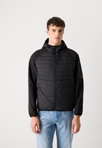 Schwarze Kapuzenjacke mit gesteppter Vorderseite, Reißverschluss und elastischen Bündchen, kombiniert mit hellblauen Jeans. Glatte Textur, minimalistischer Stil.