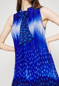 Femme portant une robe bleue sans manches avec des plis, un dégradé et un motif à pois, ainsi qu'un nœud noué au niveau du col.