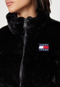 Zbliżenie osoby noszącej czarną kurtkę z sztucznego futra z wysokim kołnierzem i naszywką z logo Tommy Jeans na piersi.