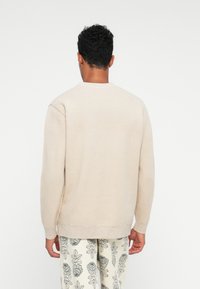 Beige kötött pulóver, kerek nyakkivágással és hosszú ujjakkal, bordázott mandzsettákkal és derékrésszel. Mintás, világos színű nadrággal viselve.