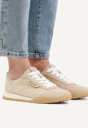 Beige Wildledersneaker mit weißen Schnürsenkeln und hellbraunen Gummisohlen, getragen mit hellblauen Jeans.