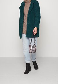 Teal gestructureerde jas over een beige coltrui, gecombineerd met lichtblauwe opgerolde jeans en zwarte haklaarzen. Houdt een gestreepte mini tas vast.