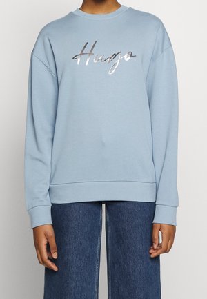 Lyseblå sweatshirt med metallisk skrift "Hugo" på brystet, båret med blå jeans, vist fra hals til hofter.