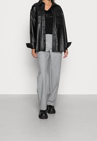 Schwarze Lederhemdjacke mit frontalen Taschen, kombiniert mit hellgrauen maßgeschneiderten Hosen und klobigen schwarzen Schuhen vor einem hellen Hintergrund.