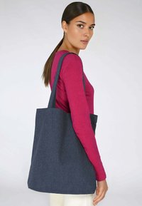 Tamna traper torba s dva čvrsta remena za rame, pravokutnog oblika, glatke teksture i minimalističkog dizajna, koju nosi osoba u ružičastoj majici.