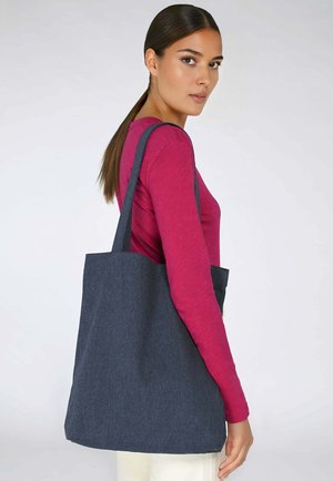 Donkerdenim draagtas met twee stevige schouderbanden, rechthoekige vorm, gladde textuur en minimalistisch ontwerp, gedragen door iemand in een roze top.