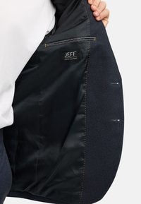 Interno di un blazer a spina di pesce navy con fodera nera liscia, cuciture gialle e un'etichetta che dice "JEFF Urban Tailoring."