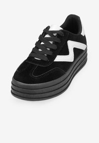 Zapatillas de ante negro con suela gruesa negra, detalles en zigzag blanco en el costado, cordones negros y lengüeta acolchada para mayor comodidad.