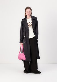 Cappotto trench nero doppiopetto sopra una t-shirt bianca con logo marrone, abbinato a pantaloni larghi neri e una borsa lucida rosa.
