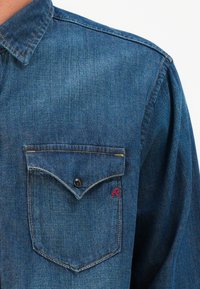 Camisa de mezclilla azul con cuello y un bolsillo abotonado. Presenta costuras en contraste y un pequeño logo bordado rojo en el bolsillo.