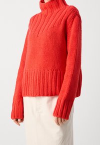 Person iført en lys rød chunky strik turtleneck sweater med lange ærmer og en creamfarvet nederdel mod en plain baggrund.