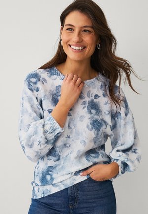 Smilende kvinde med langt brunt hår iført øreringe i ringform, en blå og hvid blomstret sweater og blå jeans, poserer med den ene hånd i lommen.