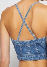 Top corto de mezclilla con tirantes cruzados en la espalda, en azul claro. Presenta detalles de costura y un pequeño logo bordado en la parte de atrás.